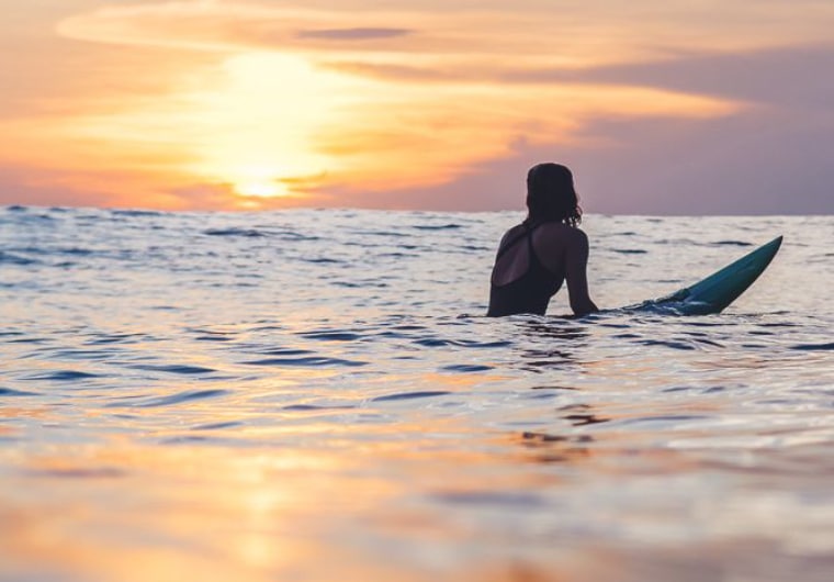 A tranquil beach setting with a woman peacefully surfing on a surfboard during a picturesque sunset.